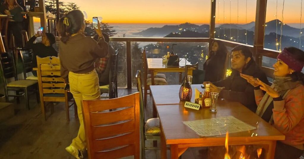 "Tourists savoring hot snacks and live music at a cozy Mussoorie hillside café, with misty mountain views and rustic wooden décor."
